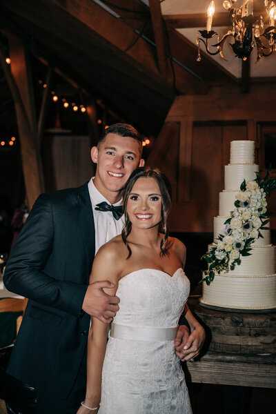 man in black tux hugging woman in white dress while standing in front of a white cake
