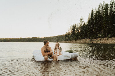 A joyful couple celebrates on a white mattress floating in a lake. The man, shirtless with a tattoo on his arm, pops a champagne bottle, causing spray to fly, while the woman, laughing and dressed in a wet white dress, sits beside him. They are surrounded by the natural beauty of a forest and the calm waters of the lake at dusk, creating a carefree and romantic atmosphere.