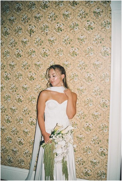 A bride smiles down her shoulder while leaning on a wallpapered wall holding her white bouquet captured by a Wilmington wedding photographer
