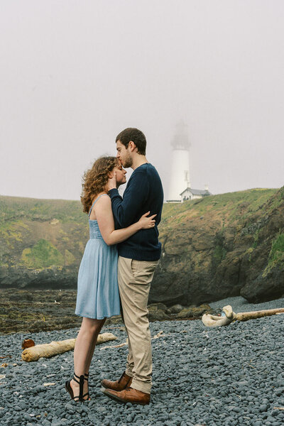 A man kisses a woman's forehead as they embrace with a lighthouse in the background. 