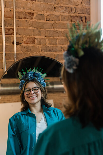 Empowerment as a woman stands before a mirror crafted in our art workshops. Adorned in a handmade wreath and a stylish jean jacket, witness the transformative journey of self-expression.