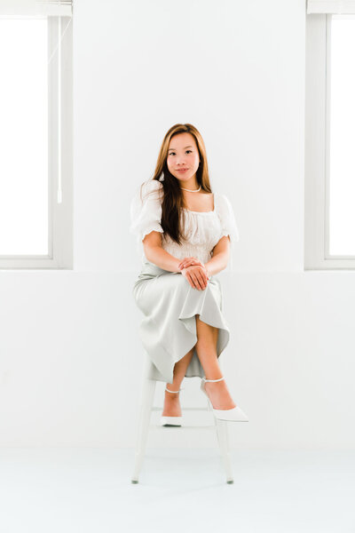 founder of Coastical Designs, sitting cross-legged on a stool in a brightly lit room, wearing white heels and a green silk skirt. This image captures the elegance and professionalism of a luxury branding designer, embodying the refined approach to high-end branding services and custom web design.