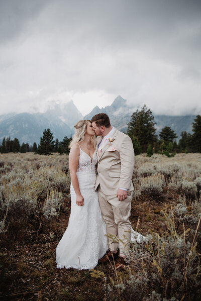 foggy wedding int he grand tetons with bride and groom kissing in a meadow with the fog rolling over the mountains