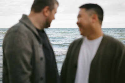 Couple smiles at each other on a beach while the lake is in focus behind them. Engagement photos by Prince Edward County photographer Jennifer van Son