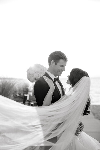 black and white image of bride and groom embracing with veil in the wind