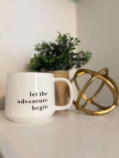 White coffee mug reading "let the adventure begin" beside greenery and gold desk decor. 