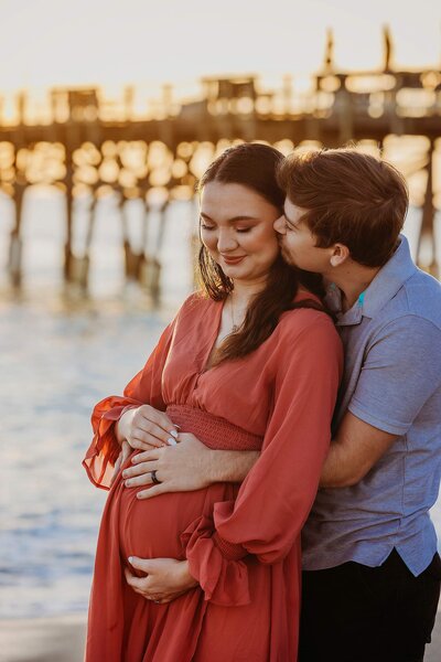 A loving pregnant couple at the beach, holding hands and gazing at the ocean, surrounded by a serene coastal atmosphere.