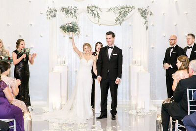 A bride and groom celebrate their just-married moment under a modern white archway at SoHo63, a Chandler wedding and event venue. Surrounded by candles, petals, and greenery, the bride raises her bouquet in triumph as the groom smiles beside her.