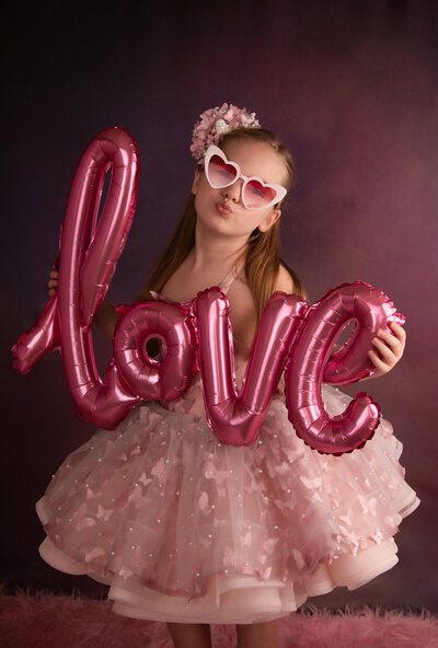girl-in-srlington-tx-studio-holding-love-balloon-and-wearing-pink-dress-with-white-heart-shapped-sunglasses