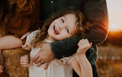 A sweet toddler girl in Fort Worth, TX smiling at the camera while her parents wrap their arms around her.