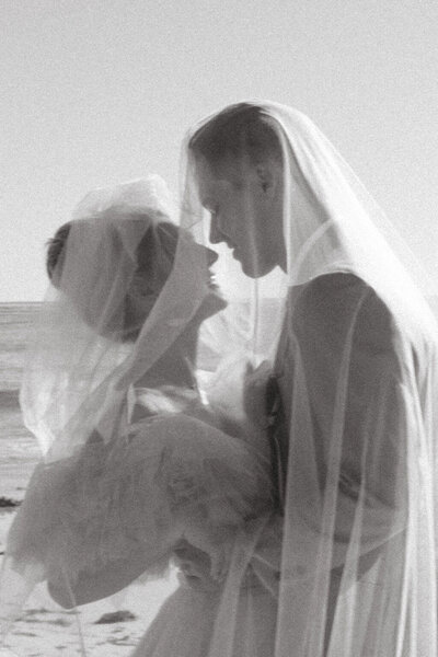 Couple in wedding attire standing in front of Carmel CA Beach under a veil almost kissing
