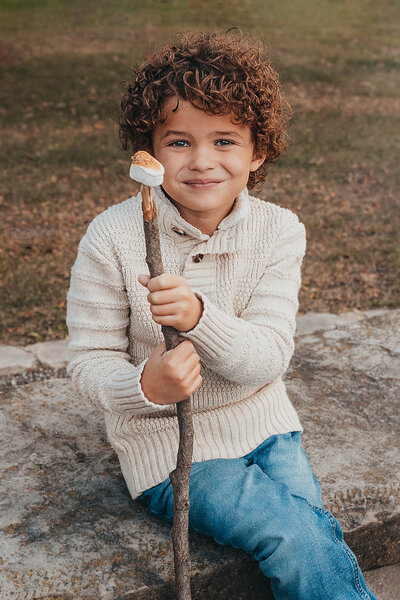Little boy sitting on stone bench. Extremely cury brown hair. He is holding on to a stick with a roasted marshmellow on the top. Boy is smiling at camera. Child Photography