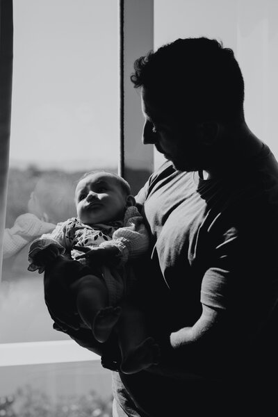 A black and white photof of a father holding his infant son in a sunny window