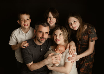 A proud father holds his newborn baby while siblings lovingly pose beside him during a dark-toned, elegant studio session by Duehr Photography in Maple Grove, Minnesota