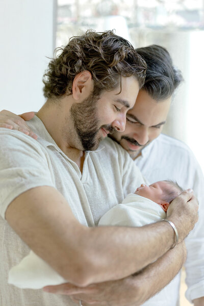 Two dads holding their newborn baby during in-home Miami newborn session by Sandra Vallejo Photography.
