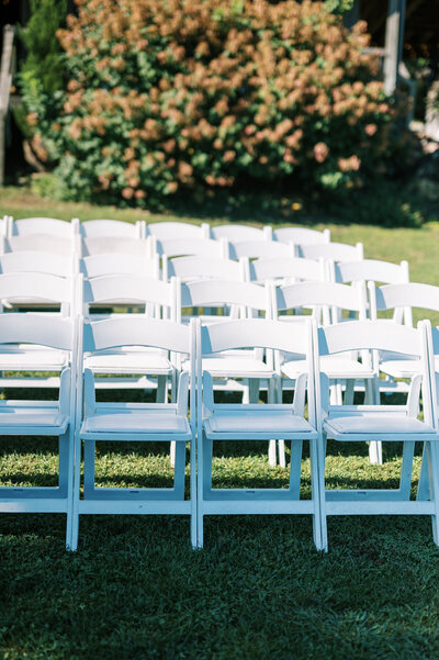 a set of white chairs for a wedding venue outside with green grass and a bush of flowers behind by Colorado Wedding Photographer JKG Photography