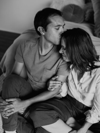 Black and white image of a wife, hugging her husband's arm as he kisses her for head during a studio photography session.