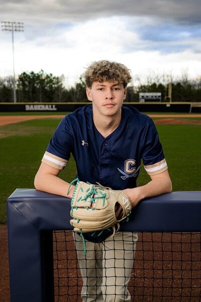 standin gon a cuthbertson baseball field a player leans against field railing holding glove and ball