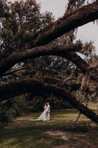 A bride and groom pose for portraits under the trees, surrounded by nature, at the Ever After Flower Barn, a wedding venue in Seffner, Florida- by Angela Moon Photography. 