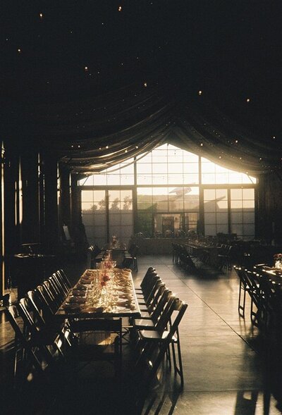 An elegant wedding reception setup inside a dark, open space with large windows letting in soft evening light. The venue is in Phoenix, Arizona, with long tables set for guests and twinkling lights strung across the ceiling.