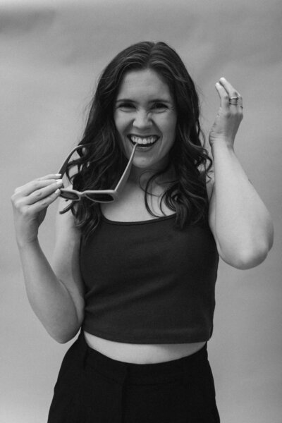A black and white photo of a girl smiling and holding sunglasses. Standing in front of a backdrop with a crop top and dress pants.