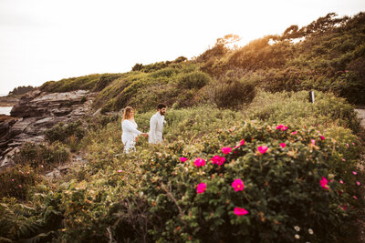 A couple walking walking through green bushes and roses on the coast of maine hand and hand.
