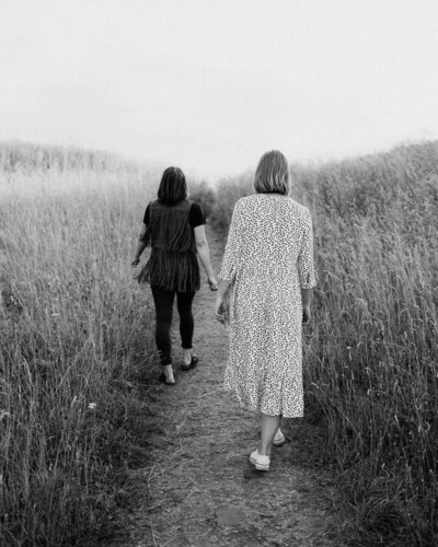 Kaksi aikuista siskoa kävelee pois päin polkua pitkin keskellä heinäniittyä Suomenlinnassa Helsingissä