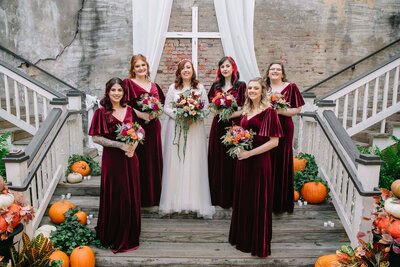A bride in a white dress stands with five bridesmaids in matching burgundy dresses, all holding bouquets, on wooden stairs decorated with pumpkins and greenery. A white cross and drapes are displayed behind them.