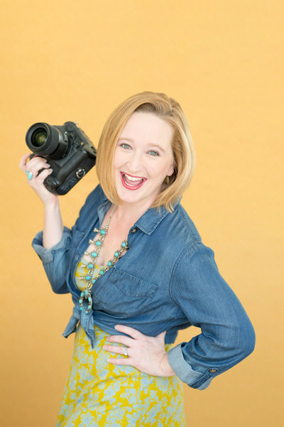 Photographer standing holding a camera with a big smile on her face and a bright yellow background in a dress and a blue jean jacket