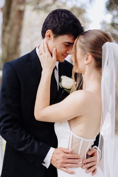 Bride and groom stand forehead to forehead, as the bride gently rests her hand on the groom's face. The bride wears pearl earrings and the groom has a white rose boutonniere. Photo taken at Disney's Grand Floridian Resort & Spa Lake Buena Vista, FL by Orlando Wedding Photographer Four Loves Photos and Film.