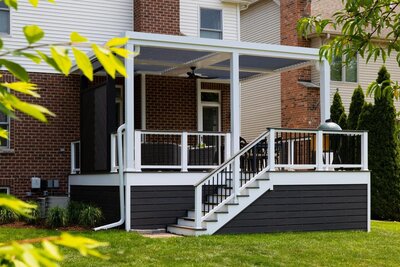 White StruXure pergola on the back of a suburban house on a deck with steps down to a green yard. 