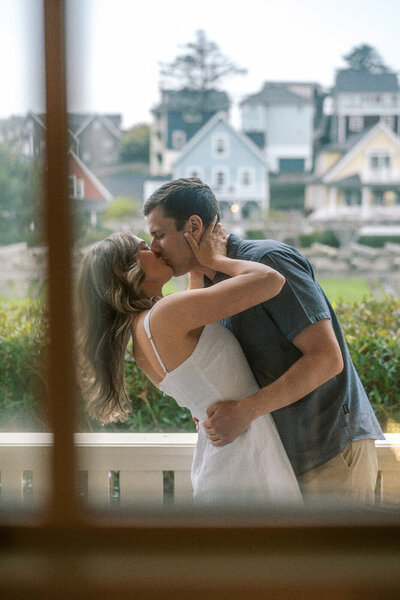 A couple are seen kissing through a window pane, representing an example engagement shoot by lifestyle photographer Among the Oaks. 