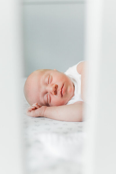 Portland Newborn Photographer_ a close up photo of a sleeping newborn baby boy in his crib. the photo is taken outside the crib so you see the crib slats in the foreground with the focus of the photo being the baby's sweet facial features and details. in home newborn photo