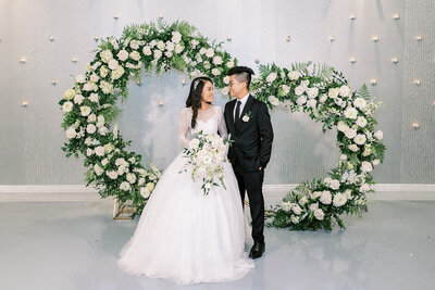 A bride and groom stand in front of a large floral heart installation at SoHo63, a Chandler wedding and event venue. The bride wears a ballgown with lace sleeves and holds a cascading white bouquet, while the groom looks at her in a classic black suit.