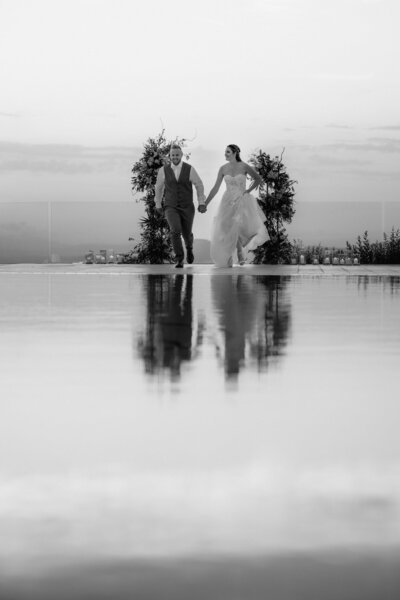 Der Anblick des in Richtung Kamera laufenden Hochzeitspaares spiegelt sich im Wasser vor ihnen wieder.