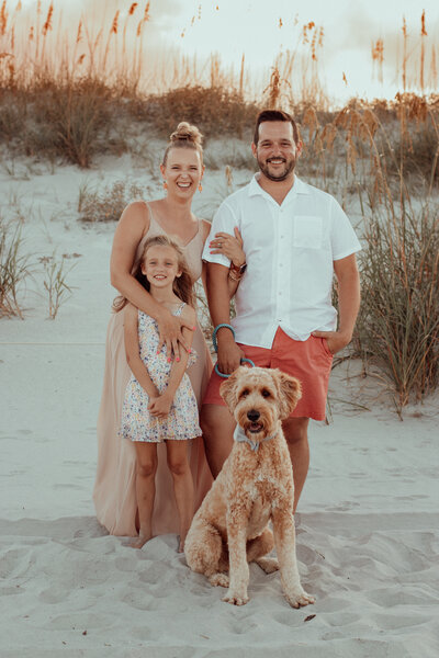 family photo of husband, wife, daughter and dog on beach