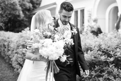 An elegant black and white photo captures the blissful moments of a bride and groom walking in front of a charming house, highlighting the timeless beauty of their destination wedding.
