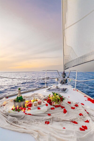 Romantic sailboat proposal setup in Hawaii with champagne, cupcakes, flowers, and rose petals arranged on the deck at sunset as the boat sails over the ocean.
