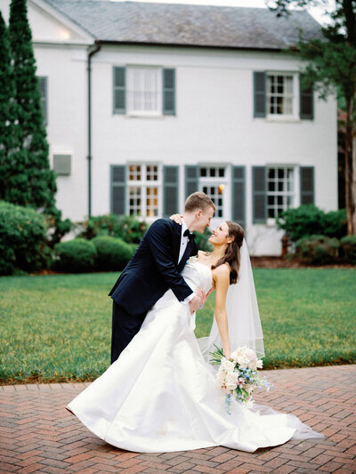 Seth and Mimi pose for their wedding photos at Boxwood Estate, surrounded by the picturesque grounds. Mimi wears a beautiful white wedding gown, and Seth is dressed in a sharp suit. The couple smiles and embraces in front of the estate’s elegant architecture and lush greenery. Soft natural light highlights the moment, creating a romantic and timeless atmosphere as they capture the joy of their special day.