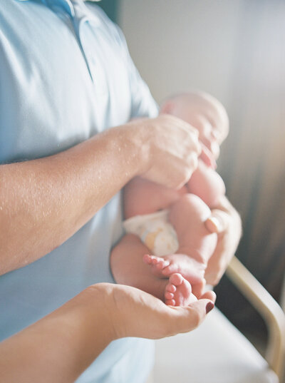 Father's arms hold naked newborn son, while mother's hands hold baby's feet