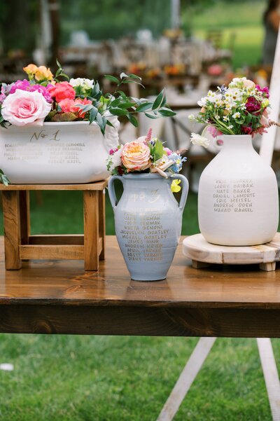Rustic wooden table with vintage ceramic vases filled with pink and white roses, set in an outdoor wedding reception.
