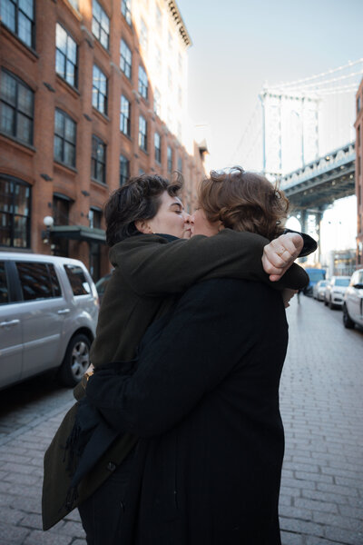 A couple embracing on a cobblestone street in DUMBO, NYC, with the Manhattan Bridge in the background.