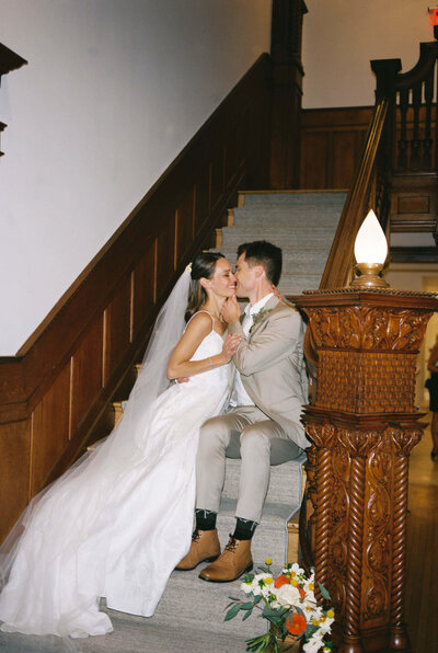 Couple in wedding attire sit on a vintage wood staircase and lean in for a kiss