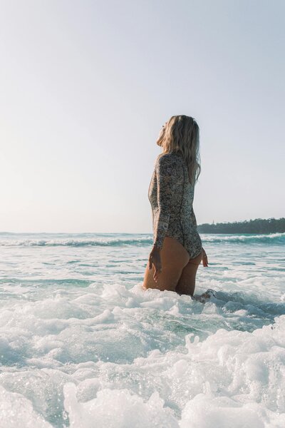 A person is captured from the back as they wade into the sea, with foamy waves around their thighs. They're dressed in a long-sleeve, patterned swimsuit that hints at a vintage style.