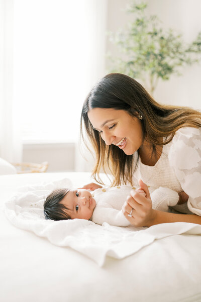 Mom in white dress hovers over dark haired baby girl who is smiling at the camera during in-home newborn photo session.
