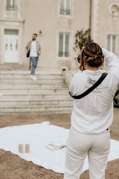 Laura de dos photographiant une scène extérieure avec un homme descendant des marches en arrière-plan, dans une tenue décontractée.