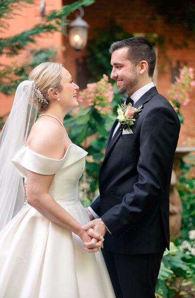 A bride in a white dress and veil holds hands with a groom in a black suit and bow tie. They are smiling at each other, surrounded by lush greenery and pink flowers. A vintage lantern hangs in the background.