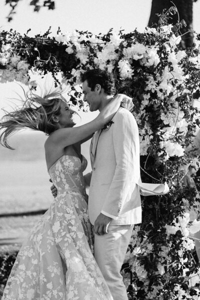 Black and white photograph of newlyweds embracing beneath a floral arch, wind blowing through the bride’s hair — timeless moment captured by Asia Pimentel