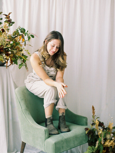 Woman in tan overalls and brown boots sitting on a green chair surrounded by florals and greenery