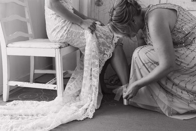 Bride's mom helps bride tie bows on her shoes while bride holds her lace gown up.